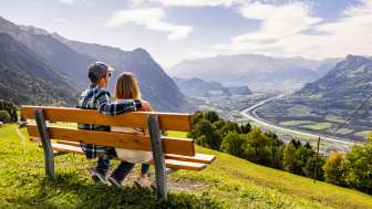 Paar sitzt auf einer Bank in Triesenberg und blickt auf das Rheintal in Liechtenstein mit beeindruckendem Alpenpanorama
