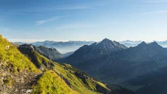 Alpiner Wanderweg mit Blick auf eine grüne Berglandschaft und schroffe Gipfel in der Morgensonne