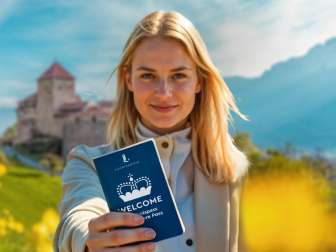 Frau hält den WELCOME Erlebnispass vor Schloss Vaduz in einem blühenden Feld hoch