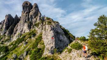 Wanderer auf felsigem Bergpfad vor zerklüftetem Grat mit rot-weissem Wegzeichen